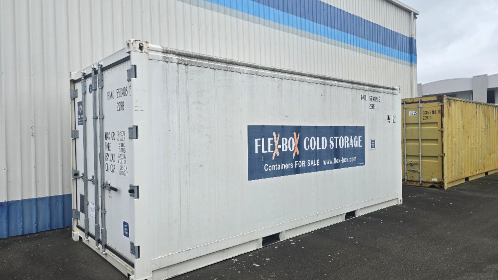 A white shipping container with a blue and white 'FLEXBOX COLD STORAGE' sign on the side, parked outdoors in front of a corrugated metal building.