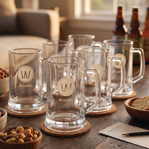 Six empty WilliamsWarn 500ml beer handle glasses displayed on a wooden table in a warm home setting.